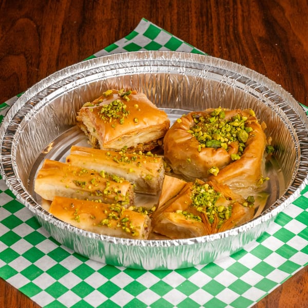 Assortment of golden-brown baklava pastries, topped with chopped pistachios, in a foil container on a checkered napkin.