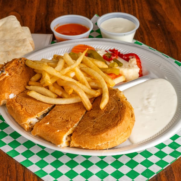Mediterranean sandwich with fries, pickled vegetables, and two dipping sauces on a white plate.