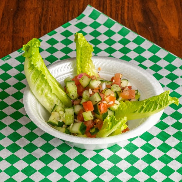 A white bowl of diced cucumbers, tomatoes, and onions, garnished with lettuce leaves on a green checkered paper.