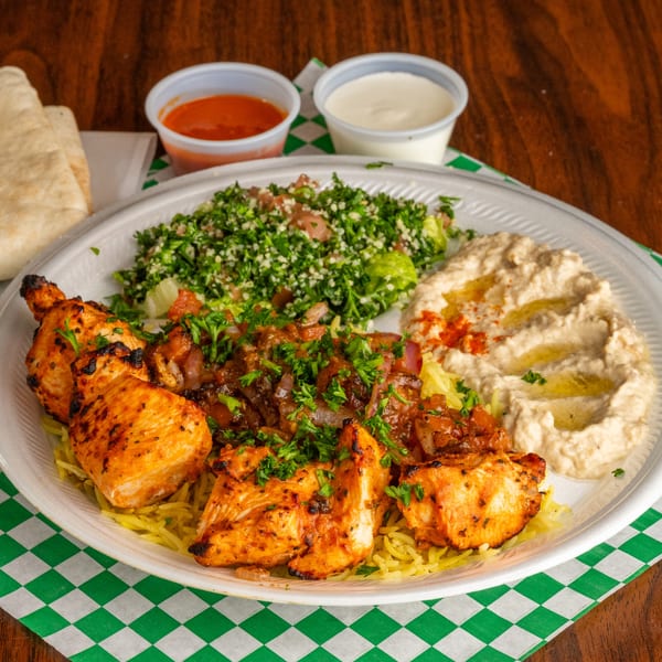 Grilled chicken skewers with rice, tabbouleh, hummus, and sauces on a white plate.