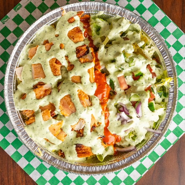 Overhead view of a Mediterranean chicken and salad platter with white and red sauces in a foil container.