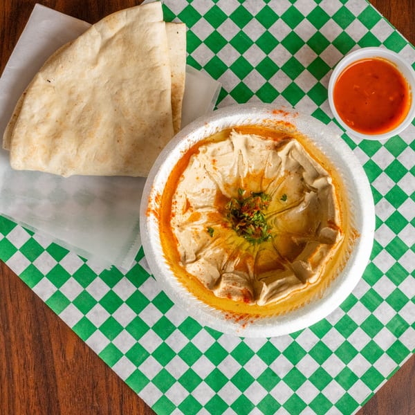 A white bowl of hummus with pita bread and red sauce on a green and white checkered paper.